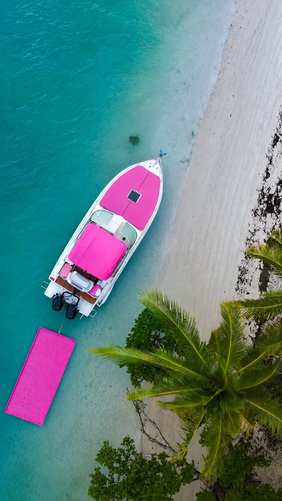 Bright pink speedboat anchored in turquoise shallow water beside a white sand beach with palm fronds visible from above